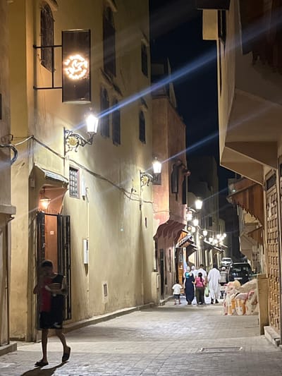 The image depicts a narrow street at night in Al Machouar – Stinia, Morocco. The street is paved with cobblestones and lined with buildings constructed from yellow and pinkish plaster. Ornate streetlights illuminate the scene, casting long shadows.
On the left side of the street, a young person wearing a red and black shirt and dark shorts walks towards the viewer, holding an object. An open doorway with a metal gate is visible behind them, with a small light illuminating the interior. Above the doorway, a decorative lamp hangs. Further up the wall, a rectangular sign with a circular, illuminated pattern is mounted.
On the right side of the street, several people are walking away from the viewer. They are dressed in traditional Moroccan attire, including a woman in a blue hijab and a long dress, a woman in a pink top and green pants, and men in white robes. A child in a white shirt and blue shorts is also present. A black vehicle is parked further down the street, and stacks of goods wrapped in plastic are piled on the sidewalk. The buildings on this side feature wooden architectural elements and ornate doorways. The overall atmosphere is dim and atmospheric, characteristic of a Moroccan medina at night.