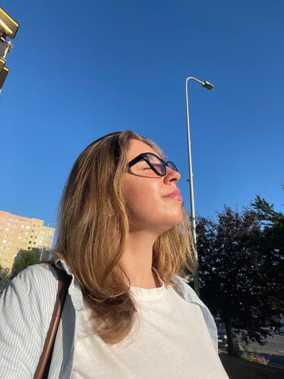 This is a close-up, vertically oriented, eye-level outdoor shot of a young person with blonde hair, wearing black-rimmed glasses and a light blue and white striped shirt over a white t-shirt. The person is looking upwards, with their head tilted back, and their eyes are closed or partially closed, suggesting they are enjoying the sun. Their lips are slightly parted and appear glossy. The sunlight is hitting their face, creating a soft glow.

The background is a clear, bright blue sky. A streetlamp with a curved pole is visible against the sky. In the distance, a dark tree with dense foliage is silhouetted against the sky, and behind that, there is a faint view of a building, possibly an apartment complex, with multiple windows. The light suggests it is daytime, likely sunny.

The subject is positioned in the foreground, with their hair and shoulders taking up a significant portion of the frame. A brown strap, possibly from a bag, is visible across their shoulder. The overall impression is one of a peaceful, sunny moment outdoors, with the subject basking in the light. There is no visible text in the image.