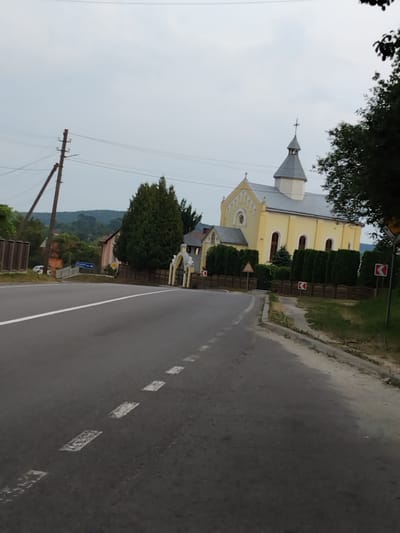 The image depicts a scene along a road in Khodorkivtsi, Ukraine. The road has a white dashed line marking its center and a light-colored shoulder. On the left side of the road, there is a yellow church with arched windows and a grey steeple topped with a cross. The church is surrounded by a hedge and a fence. Beyond the church, lush green trees and rolling hills are visible, suggesting a rural or semi-rural setting. A wooden utility pole with wires is also present on the left side of the road. The sky is overcast, indicating a cloudy day, and the lighting suggests it is either early morning or late afternoon. There are no people visible in the image, and the overall atmosphere appears calm and serene. Several road signs with red and white markings are visible along the roadside. One sign is a blue directional sign with white text.