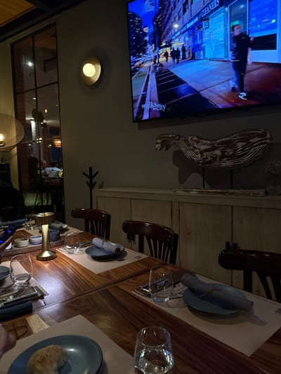 A sideways view captures a restaurant scene. A TV displays city scenes next to a decorative whale. Tables are neatly set with plates, glassware, and silverware, arranged on placemats. Chairs are positioned around the table. A light fixture illuminates the scene. Further back, a partially visible reflection shows the restaurant's interior.
