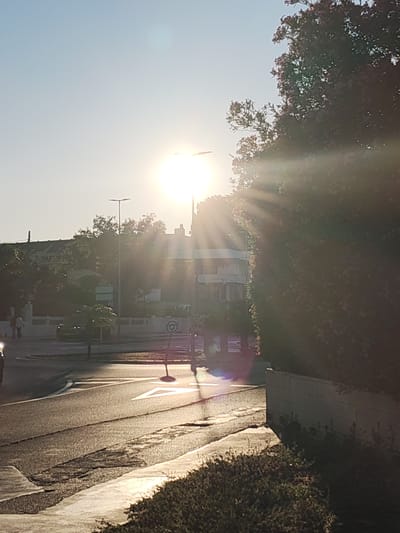 The image depicts a street scene in Rotes, Spain, bathed in bright sunlight. A road with marked crosswalks and a roundabout is visible. A street lamp stands near the roundabout. Buildings and trees line the street in the background. Two figures can be discerned walking in the distance. A car is visible on the left side of the frame. Lush greenery is present on the right side of the image, along a wall. The sun is high in the sky, creating intense glare and rays of light.