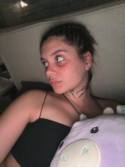 The image shows a close-up, low-angle portrait of a young woman with dark hair pulled back, wearing a black spaghetti strap top. She has multiple piercings, including a septum ring and an earring. There are tattoos on her neck, one appearing to be a spider. She is holding a purple stuffed animal, possibly a cow or dragon, with visible eyes and ears. The woman is looking to her left, with rosy cheeks and her hair slightly disheveled. The background appears to be indoors, with a textured surface and some lighting that suggests a dimly lit environment. There is no visible indication of the city of Genemuiden, Netherlands, in this image.