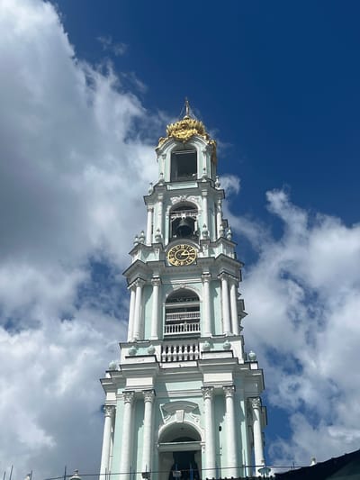 This is a low-angle, eye-level shot of the Bell Tower of the Trinity Lavra of St. Sergius in Sergiyev Posad, Russia. The tower is a pale teal with white architectural details and a golden crown at the very top. A large, round clock with Roman numerals is visible on one side of the tower, showing approximately 9:57. A bell hangs within an arched opening above the clock. The tower is set against a vibrant blue sky with dramatic, white cumulus clouds. Some construction scaffolding is visible at the very bottom of the frame.