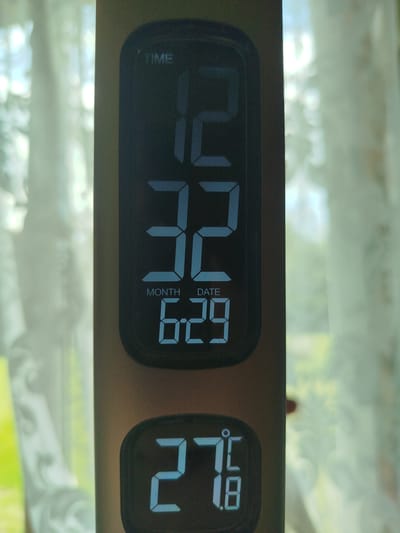 The image displays a digital clock and thermometer. The top display shows "TIME" with the numbers "12" and "32" prominently featured. Below this, it indicates "MONTH" and "DATE" with the numbers "6" and "29". The bottom display shows a temperature reading of "27.8" with a degree symbol. The device is held vertically. The background is blurred and appears to be an indoor setting with light filtering through a window covered by sheer curtains, suggesting natural light. The overall impression is of a modern electronic device set against a soft, natural backdrop.