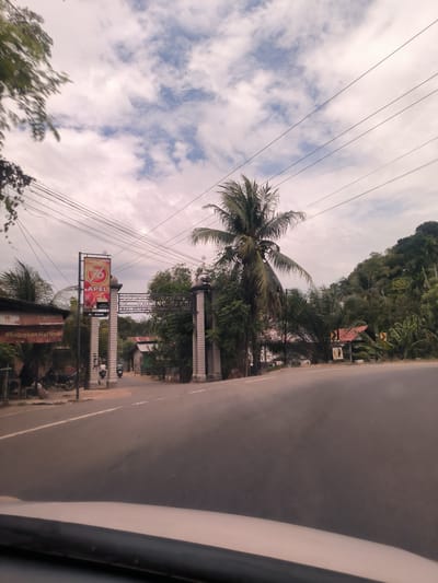The image captures a rural or semi-rural scene in Sawang, Indonesia, taken from inside a vehicle moving along a winding asphalt road. The road curves to the right, leading towards a hill densely covered with tropical foliage, including prominent palm trees and what appear to be banana plants. On the right side of the road, there's a small sign with a red roof and black and yellow chevron arrows, indicating the curve ahead.
On the left side of the road, a decorative gateway with two tall, ornate columns marks the entrance to a village or settlement. Above the entrance, a sign clearly reads "SELAMAT DATANG DI GAMPONG CARO KEC. PEUDADA KAB. BIREUEN," which translates to "Welcome to Gampong Caro, Peudada District, Bireuen Regency." Next to the gate, a tall billboard advertises "DIARIUM BARU" and "76 APEL" with "AL-SAHSI" written below. Below this billboard, another sign displays "#Extrakam Harimu" (likely a marketing slogan).
Several modest buildings with corrugated metal roofs are visible behind the gate and further along the left side. Two individuals on motorbikes are seen riding through the gateway, moving away from the camera. Overhead, numerous power lines stretch across the sky, crisscrossing the frame. The sky is partly cloudy, with patches of blue visible between white and grey clouds, suggesting it is daytime. The overall impression is of a quiet, tropical setting.