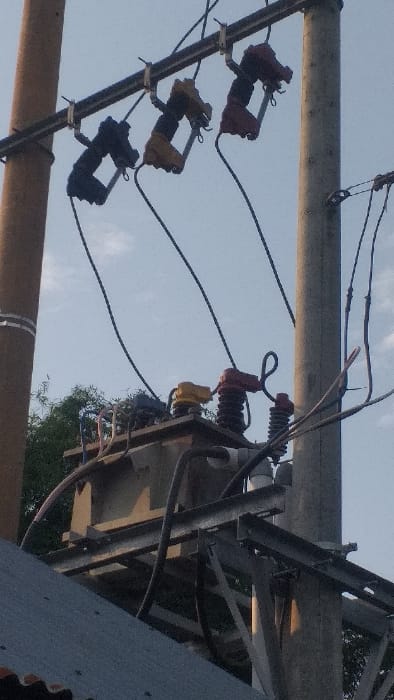 This image depicts electrical infrastructure on a clear day in Lamreung, Indonesia. Two utility poles are visible: a brown pole on the left and a gray concrete pole on the right. A metal crossarm is mounted between the poles, supporting three overhead primary cut-out fuses, colored black, yellow, and red from left to right. Electrical wires extend downwards from these fuses. Below the fuses, a large gray electrical transformer is mounted on a metal support structure positioned between the two poles. The transformer has various colored insulators (yellow and red are prominent) and multiple electrical cables connecting to and from it. Additional wires and cables are visible, running between the poles and the transformer. In the background, green tree foliage is present under a light blue sky with sparse white clouds. The corrugated dark gray roof of a building is visible in the lower left foreground. No individuals are present in the scene, and no active human actions or interactions are depicted.