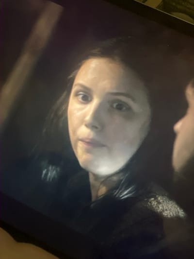 A woman with dark hair and fair skin gazes at the viewer. The image is dark, possibly from a screen capture, with a head partially obscuring the top of the frame. The woman's expression is neutral, and her face is well-lit compared to the background. She wears dark clothing, and her hair appears sleek.