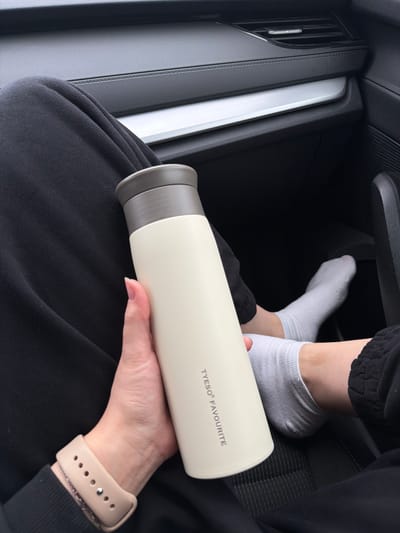 The photograph depicts the interior of a car. An individual is holding a white TYESO Favourite thermos in their hands, which are adorned with a light beige smartwatch. The individual is wearing black clothing, likely sweatpants. In the background, a pair of feet wearing white socks are visible near the dashboard area. The left side of the image shows part of the car's interior, including a silver trim and an air vent. The photograph appears to be taken indoors. It is unknown if the photograph was taken in Fedotovo, Russia.
