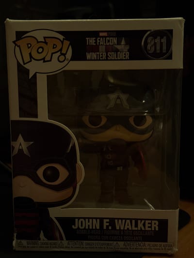 A close-up, dark, eye-level shot captures a Funko Pop figure of John F. Walker from "The Falcon and the Winter Soldier" inside its clear plastic packaging. The figure, dressed in a red and dark blue costume with a star emblem on its chest, has a dark mask covering its face and large, round yellow eyes. The packaging displays the Marvel Studios logo, the show's title, and the number "811." The bottom of the packaging includes safety warnings in English, French, and Spanish, indicating it is a bobble-head figure and not suitable for children under 36 months. The background is dark and out of focus. There is no discernible location in Zhovte, Ukraine.