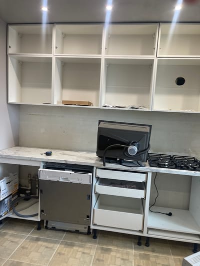 The image depicts the interior of an unfinished kitchen in Rechytsa, Belarus. A row of white upper kitchen cabinets are installed, some containing boxes and plastic wrap. Below the cabinets, a white countertop with a marble-like pattern is installed. On the countertop, a dark gray sink is positioned upside down, with plumbing fixtures attached. To the right of the sink is a black gas stovetop. Beneath the countertop, a dishwasher is partially installed, with its door open. To the right of the dishwasher are three white drawers, two of which are open, revealing their contents. A black electrical cord with a plug is visible on the floor to the right. The floor is tiled with square, beige tiles. The walls appear to be unfinished, with visible drywall. The lighting is provided by ceiling spotlights. There are no people present in the image.