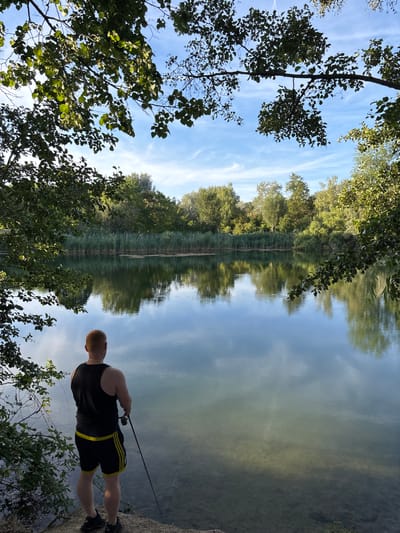 A man with red hair is standing on the bank of a lake in a park, fishing. He is wearing a black tank top and black shorts with yellow stripes. The lake is surrounded by trees and reeds. The sky is blue with some clouds. The scene is peaceful and serene. This lake is located in the city of Ludwigshafen am Rhein, Germany.