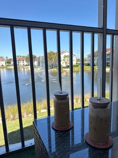 The image captures a static scene from a residential balcony in a condominium community in Naples, Florida, United States. Two clear glass can-shaped cups, each containing an iced, light-brown beverage, rest on a dark, woven-textured outdoor table with a reflective glass surface. Each cup is topped with a circular wooden lid and a clear glass straw, and positioned on a round, reddish-brown coaster. The balcony is enclosed by a dark metal railing with vertical balusters and a fine mesh screen. Beyond the railing, a man-made lake or pond spans the midground, featuring an active fountain spraying water in a broad, fan-like pattern. The water's edge is lined with light green vegetation. In the background, multiple multi-story residential buildings with light-colored facades and reddish-brown tiled roofs are visible, interspersed with trees. A utility pole or tower is faintly visible in the far distance. The sky is clear and blue. No individuals are present in the frame.