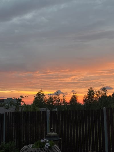 The image depicts a sunset over a residential area. In the foreground, a dark wooden fence stretches horizontally across the frame, with a woven basket containing white flowers and a lamppost to its right. Beyond the fence, a line of trees with green foliage forms a natural barrier. In the background, buildings and rooftops are visible, suggesting an urban or suburban setting. The sky is filled with clouds, with hues of orange, yellow, and gray indicating the time of day is sunset. The overall scene is peaceful and serene. The location is the city of Gubakha, Russia.