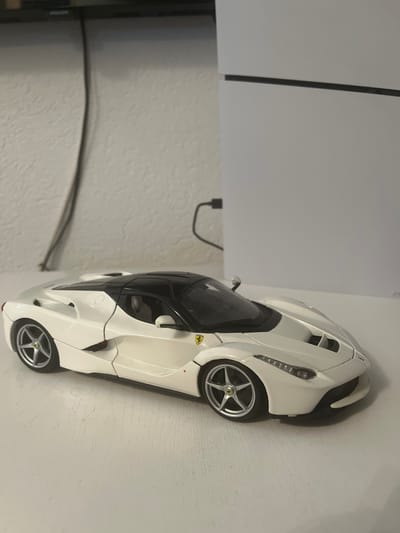 A white Ferrari toy car is positioned on a white surface, possibly a table. The car is facing right, with its front slightly angled towards the camera. The background shows a textured white wall with a black cable running down it and a large white rectangular object, possibly a game console or appliance. A black cable is also connected to the white object. The overall lighting suggests an indoor setting. No people are present in the image. The location in Neustadt, Germany, is not discernible from the visual information provided.