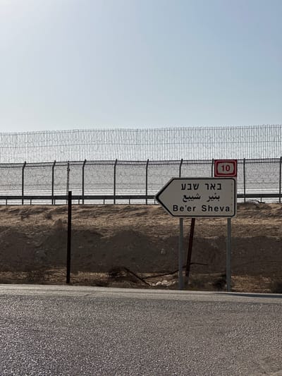 The image depicts an arid, desert landscape under a clear, bright sky. A tall security fence, constructed from chain-link mesh and topped with multiple strands of coiled razor wire, extends horizontally across the middle ground. Behind the fence, a raised, light brown sandy embankment is visible, partially obscuring a track or road. In front of the fence, a similar sandy slope descends towards a paved asphalt road that occupies the lower third of the frame.
A white, arrow-shaped directional sign is positioned in the lower right-midground, pointing left. The sign bears the destination "Be'er Sheva" written in Hebrew (באר שבע), Arabic (بئير شيبع), and English. Above this sign and affixed to a fence post is a smaller red rectangular sign with a white border, displaying the number "10" in white, indicating Route 10. Several metal posts support the signage and are visible along the roadside and fence line. The environment is devoid of people or discernible activity.
The scene is located within the Ramat Negev Regional Council, Israel, specifically along Route 10 in the Negev Desert, near the border with Egypt, as indicated by the "Be'er Sheva" sign and the prominent Route 10 marker.