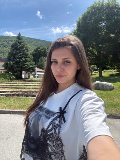 The image is a bright, sunlit selfie taken outdoors, featuring a young woman against a natural and rural backdrop. The photograph is rotated 90 degrees clockwise, presenting the scene sideways.
The primary subject is a young woman, shown from the chest up. She has long, straight, dark brown hair, a fair complexion, and a gentle smile. Her dark eyes are directed towards the camera. She is wearing a white t-shirt that features a prominent black lace pattern across the chest and shoulder area, accompanied by a small black ribbon or bow near the left shoulder.
The setting is a picturesque outdoor environment. The sky is a clear, vibrant blue with a few scattered white clouds, suggesting a sunny daytime. In the background, a large, green, tree-covered mountain or hill dominates the horizon. Various types of trees, including large deciduous trees with full green canopies and darker coniferous trees, are visible throughout the mid-ground and background.
To the woman's right, there is a paved path or road next to a grassy area, where a large, light grey rock is situated. To her left, behind some foliage, part of a building with a red roof can be seen. Below this building, there are distinct terraced structures or steps, possibly for agricultural or landscaping purposes, extending across the mid-ground. The overall impression is one of peace and natural beauty. There is no discernible text visible in the image. The location context indicates this scene is in Vetren Dol, Bulgaria.