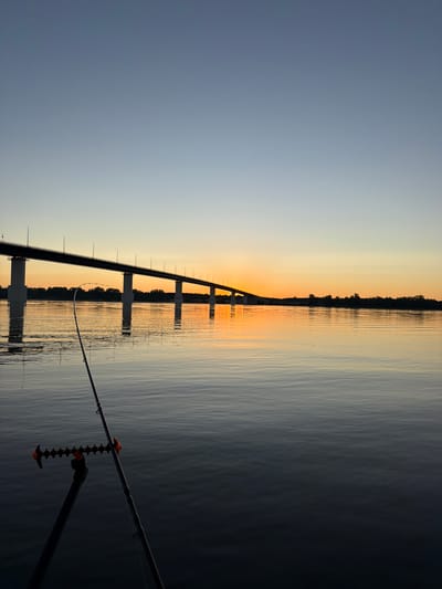 A fishing rod is set up on a tripod by a body of water. The rod points towards the right side of the frame. In the background, a long bridge spans across the water. The sky is clear with a gradient from light blue at the top to a warm orange glow near the horizon, indicating either sunrise or sunset. The water is calm and reflects the sky and the bridge. Trees and vegetation line the distant shore. The scene is serene and peaceful, capturing a moment of angling during twilight. The location is the Kama River near Kambarka, Russia.