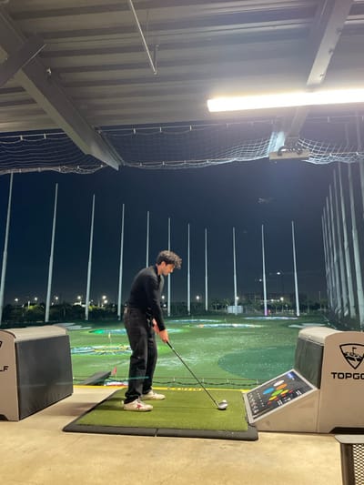 A man in dark clothing is playing golf at night at a Topgolf venue. He stands on a green mat, holding a golf club, and is in the middle of his swing. Artificial lighting illuminates the scene. In the background, illuminated targets are visible on a dark green golf course. Tall, evenly spaced poles stretch upwards. A Topgolf scoring system is positioned beside the man. The structural elements of the venue, like metal beams and netting, are visible on the left side.