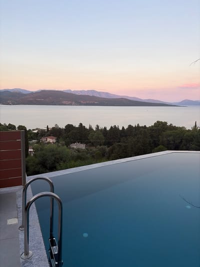This is a vertical, slightly tilted shot of a tranquil scene at dusk by an infinity pool. The pool's serene, deep blue water fills the right side of the frame, reflecting the sky and a thin, leafless branch. A sleek, silver pool ladder with two curved handles leads into the water from the bottom right corner, resting on a textured gray surface. Beyond the pool, a lush green landscape of trees and vegetation slopes down towards a calm body of water. In the distance, hazy mountains rise, their peaks softened by the fading light. The sky transitions from a pale, soft blue at the top to warm hues of pink and orange near the horizon, indicating either sunrise or sunset. A few scattered structures with red-tiled roofs are visible nestled within the trees. The overall atmosphere is one of peace and relaxation, enhanced by the soft, diffused light. There is no visible text.