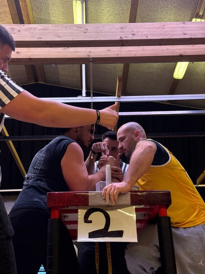 This image depicts an arm wrestling match in an indoor setting, likely a gymnasium or hall, in Reisenberg, Austria. Two male competitors are actively engaged at a red and white striped arm wrestling table, which features a metal pin handle on each side. A white sign with a large black number "2" is affixed to the front of the table.
On the left, a competitor with dark hair and a beard wears a black sleeveless shirt and dark pants. His right arm is on the table, locked with the opponent's. On the right, a bald competitor wears a yellow and blue sleeveless shirt and grey sweatpants. His left arm is similarly engaged on the table.
A referee or official, wearing a black and white striped shirt, stands between the two competitors, actively gripping their joined hands at the center of the table, appearing to ensure correct starting position or control the match. Another official, partially visible on the far left, also wearing a black and white striped shirt, extends his right arm forward, displaying a thumbs-up gesture. He wears a black wristband with "VIP" text on it.
The background reveals a high ceiling constructed with wooden beams and lighter-colored panels. Several fluorescent tube lights illuminate the scene. A dark fabric or wall covers the rear. A white banner or strap with faint text, possibly "WAF WOF," is visible below the "2" sign on the table.
