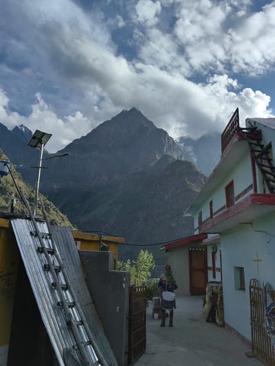 In Tiling, India, a person carries a bucket while walking down a pathway. The person is wearing a multi-colored sweater, a green scarf, and brown pants. They are walking towards a light blue and white house with red trim. A wooden ladder is leaning against the side of the house. In the background, a large mountain looms under a cloudy sky. A solar panel is mounted on a pole to the left of the frame. The scene appears to be a residential area in a mountainous region.