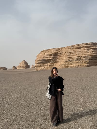 The image depicts a person lying horizontally on a gravel-covered ground near a cliff. The person is wearing a dark jacket and brown pants. A shiny bag is also visible. The background features a series of tall, brown, ridged cliffs under a grey sky. This scene is consistent with the Yadan National Geological Park near Dunhuang City, China.