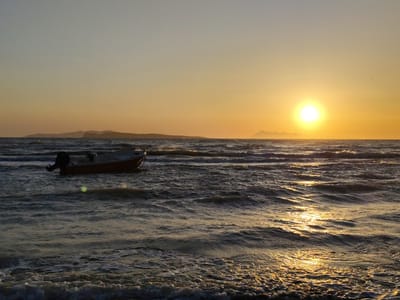 This image captures a coastal scene at either sunrise or sunset, with the location identified as Agios Stefanos, Greece.
The sky occupies the upper third of the frame, displaying a gradient from a pale, light blue-grey at the top to a warm, bright orange and yellow directly above the horizon. A prominent, intensely bright, golden-yellow sun is positioned low on the horizon, slightly to the right of the frame's vertical center. A faint, linear white contrail from an aircraft is visible in the upper left quadrant of the sky. A small, dark silhouette of a bird is present in the upper right quadrant, near the sun.
Across the horizon, a series of dark, undulating landmasses, appearing as distant mountains or hills, stretch from left to right, partially obscured by atmospheric haze.
The lower two-thirds of the image depict the sea, characterized by choppy, undulating waves with visible white foam crests. A strong, shimmering path of golden sunlight reflects on the water's surface, extending from the sun towards the foreground, particularly concentrated on the right side of the frame. The water itself appears dark blue-grey, contrasting with the bright reflections.
In the middle-left section of the water, a single, small motorboat is present. The boat has a dark hull, appearing reddish-brown under the lighting, and a visible black outboard motor at its stern. A thin, dark mooring rope is faintly discernible attached to the boat's bow. A distinct, circular greenish-yellow lens flare is visible on the water's surface to the left of the boat. No people are visible in the scene. The overall environment is marine and exposed, with dynamic water movement under intense natural light.