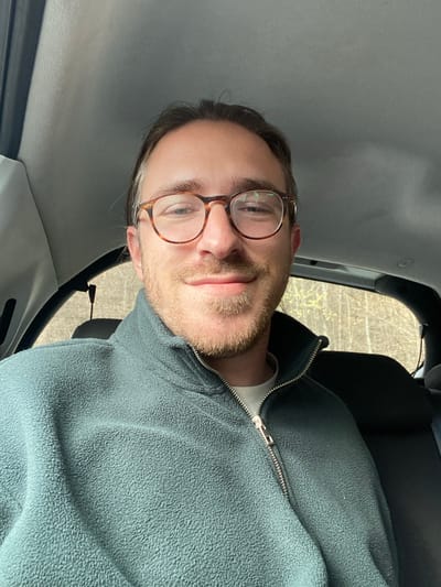 The image depicts an adult male taking a selfie while seated in a vehicle, located in the city of Kolašin, Montenegro. The male subject has light skin, a short beard, dark hair pulled back, and wears tortoiseshell-framed eyeglasses. He is dressed in a dark teal fleece jacket with a partially unzipped silver zipper, worn over a light-colored undershirt, and displays a faint smile. The vehicle's interior features a light grey headliner and dark fabric seating visible in the background. Through the vehicle's windows, a natural outdoor environment with trees displaying light green foliage is visible behind the subject.