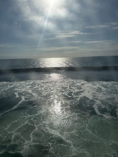 This image captures a dynamic seascape under a partly cloudy sky. The sun is high in the sky, casting bright reflections on the water's surface and creating a strong lens flare. The ocean exhibits a range of textures, from the smooth expanse of the horizon to the turbulent, foamy churn of waves closer to the foreground. The water color shifts between deep blues and greens, with white foam highlighting the movement of the waves. There are no people, structures, or specific landmarks visible to indicate a particular location within the city of Unknown, Denmark, or any other identifiable place. The scene is purely natural, focusing on the interplay of light, sky, and sea.