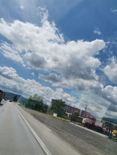 The image captures a roadside view from within a vehicle, likely on a highway. The sky is predominantly blue, filled with large, white, fluffy clouds. In the foreground, a multi-lane asphalt road stretches into the distance, marked with double white lines. On the left side of the road, several vehicles are visible, including a large truck and cars. To the right of the road, there is a gravel area adjacent to a fence. Beyond the fence, a red tractor is prominently displayed. Further in the background, there are buildings with a modern architectural style, trees, and distant mountains. Power lines and electrical towers are also scattered throughout the scene. The overall impression is that of a bright, partly cloudy day along a major road.