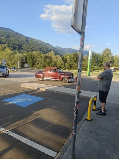 A person stands by a yellow bollard on the right side of the image, facing left. They are wearing a gray t-shirt, dark shorts, and dark athletic shoes. In the middle ground, a vintage red convertible is parked. A light-colored modern car is parked to the left of the convertible. A blue handicapped parking symbol is painted on the asphalt in the foreground. The scene is set outdoors, with a mountainous, tree-covered landscape in the background under a blue sky with scattered clouds. A signpost with stickers is prominently positioned in the foreground, slightly off-center to the right. The ground appears to be a paved surface, possibly a parking lot. The lighting suggests it is late afternoon or early evening, with long shadows cast across the ground.