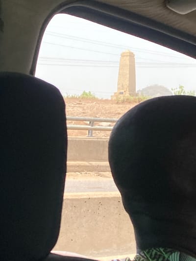 Captured from within a vehicle, the image features a tall, light-brown, brick-like tower visible through the window. The tower is situated on a dry, hilly landscape with some vegetation. An electric line runs through the sky. In the foreground, parts of the car interior are prominent, including the window frame and dark-colored seat headrests, partially obscuring the view. A fence is also visible on the side.