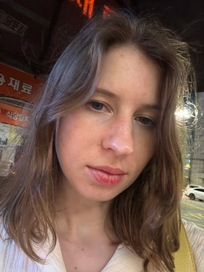A close-up, selfie-style portrait of a young Caucasian woman with shoulder-length, light brown hair. She is wearing a white, textured shirt. The background appears to be an urban setting at night, with visible Korean signage, possibly from a restaurant or shop. The lighting suggests artificial light sources, including a bright, circular light fixture to her right. A street with a car is visible in the far background.