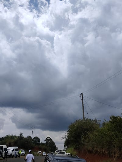 A cloudy sky dominates the scene, partially revealing blue sky patches above. Below, vehicles line a road with pedestrians nearby. Green bushes and trees flank the road. Utility poles with power lines stretch across the foreground, adding a touch of human infrastructure to the natural setting. The dark clouds suggest an approaching storm.