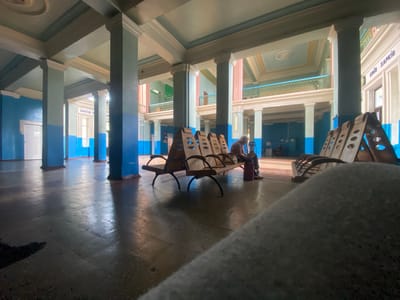 The image depicts the interior of a building that appears to be a public waiting area, possibly a train station, in Romodan, Ukraine. The architecture features tall, blue and light green painted columns supporting a high ceiling with ornate molding. Balconies with railings are visible on upper levels. A long row of empty wooden benches with metal legs is arranged in the foreground. A single individual, an elderly person with grey hair, is seated on one of the benches, wearing a reddish-brown top and dark pants, with a red bag placed beside them. The floor is tiled and reflects the light. The overall impression is one of a somewhat dated but grand public space.