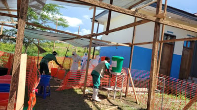 This image depicts an outdoor sanitation or decontamination station, likely associated with a health facility or emergency response in Tchere, Democratic Republic of the Congo. The scene unfolds under a bright, clear daylight sky.

In the foreground, a makeshift structure made of wooden beams and a light blue tarp provides shade over an area partially enclosed by orange plastic mesh fencing. Two men in green uniforms are actively involved in sanitation tasks. One man, wearing a beige vest and white boots, is washing his hands at a green water dispenser on a wooden stand. A label on the dispenser reads "...hloré 0.05%", suggesting a chlorine solution. To his left, another man in a dark green uniform, face visor, and gloves is bending over multiple plastic buckets, including orange ones. One orange bucket clearly displays "CLIMA 0.5%".

Behind these individuals, within the fenced perimeter, three people are seen wearing full white personal protective equipment (PPE) hazmat suits, complete with hoods, goggles, and gloves. They appear to be handling long poles or tools, indicating procedures related to disease control or hazardous material management.

In the background, a permanent building with a white upper facade and a blue lower section is visible. Wooden doors and windows mark its structure. French text on the building reads "IMAGERIE" (Imaging) and partially "CHAMBRE DE COMMANDE" (Control Room), suggesting it is part of a medical or hospital complex. Beyond the station, the natural landscape includes green trees, hills, and open scrubland under a blue sky with some clouds. The overall scene portrays a diligent public health operation focused on hygiene and safety protocols. The watermark "Joseph Kisuki" is present in the lower right.