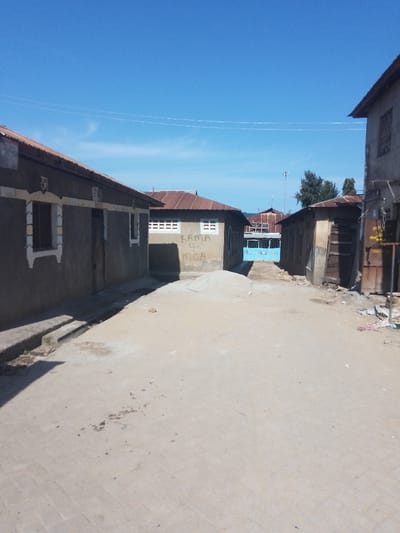 The image displays an unpopulated urban pathway under a clear blue sky in Ziwa La Ng'ombe ward, Kenya. The foreground consists of light-colored, possibly concrete paver, ground, leading to a central mound of light-colored sand or gravel in the mid-ground. On the left, two single-story buildings are visible. The nearest features a grey-brown stucco facade, a rusted corrugated metal roof, a white-framed recessed window with decorative trim, and a dark, rectangular doorway further along its wall; a drainage channel is present at its base. Behind it, a similar building displays horizontal white-framed vents and black graffiti text reading "RAMA 4 MCA". On the right, a darker, two-story building with wooden panels/doors and accumulated debris, including discarded items, is present in the foreground. Further back on the right, another single-story building with a dark facade and corrugated roof is visible. In the mid-ground, a prominent light-blue vehicle or structure, bearing white text (partially legible as "MSOMBE"), is positioned at the pathway's end. The distant background reveals additional buildings with corrugated roofs and sparse green foliage. Two thin utility lines traverse the upper sky. No individuals are present, and no actions or interactions are depicted.