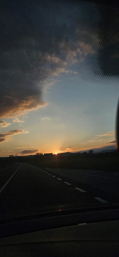 This image depicts a sunset over a rural or semi-rural landscape in the vicinity of Dolna Banya, Bulgaria, viewed from the interior of a moving vehicle. The scene is bisected horizontally by the horizon line, with the sky occupying the upper two-thirds and the road and vehicle interior the lower third.
The sky exhibits a dramatic transition from dark, heavy cumulonimbus clouds in the upper left to a clear, pale blue expanse in the upper right. Scattered across the middle of the sky are altocumulus clouds, illuminated by the setting sun with hues of orange, pink, and gold. A distinct, thin white contrail from an aircraft is visible in the clear blue portion of the sky in the upper right. The sun is prominently featured near the horizon in the mid-left, partially obscured by distant landforms, emitting bright orange-yellow light and distinct crepuscular rays that fan upwards.
The horizon consists of a continuous line of silhouetted mountains or hills, appearing dark against the bright sky. Indistinguishable structures or dense vegetation are silhouetted directly beneath the sun.
The foreground is dominated by a dark asphalt road extending from the bottom center into the distance. It features a solid white line on its left edge and broken white dashed lines marking the right lane. The immediate right side of the road is dark and indistinct, suggesting undeveloped land or sparse vegetation. The bottom section of the image shows the dark, underexposed interior of a vehicle, likely the dashboard and a portion of the windshield frame. A visible grid pattern, characteristic of a heated windshield or defroster lines, is present in the upper right quadrant, indicating the photo was taken through glass. No people are visible in the image.