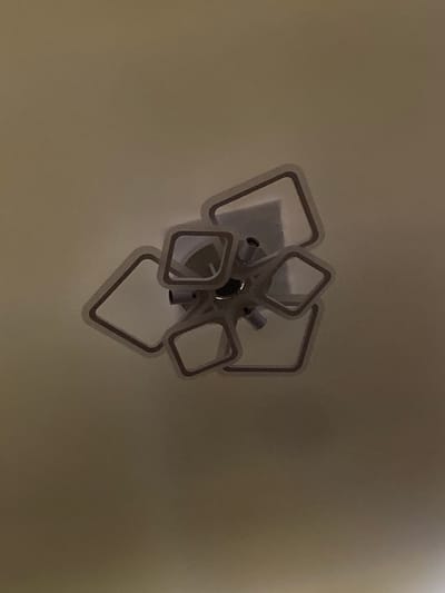 The image displays a ceiling light fixture with a modern, abstract design. The fixture consists of several square-shaped frames arranged in a cluster, emanating from a central point on the ceiling. The frames are outlined in a darker color, possibly brown or black, against a lighter interior. The lighting in the image is dim, casting subtle shadows and giving the fixture a slightly hazy appearance. The ceiling itself is a uniform, pale color, likely white or cream. The perspective is from below, looking directly up at the fixture. There are no people or any other objects visible in the frame. The location is identified as Badalbay, Uzbekistan.