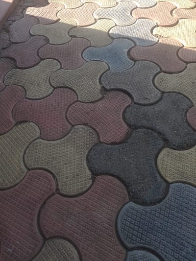 The image shows a close-up view of a patterned sidewalk in Ganja, Azerbaijan. The sidewalk is made of interlocking concrete pavers shaped like three-leaf clovers, arranged in a repeating pattern. The pavers are various shades of red, brown, grey, and yellow. The pavers are slightly uneven and have a textured surface. There are no people or other objects present in the image. The image appears to have been taken on a sunny day as there are shadows cast across the sidewalk. The location of the image is a sidewalk in Ganja, Azerbaijan at the coordinates 40.688 latitude and 46.388 longitude.