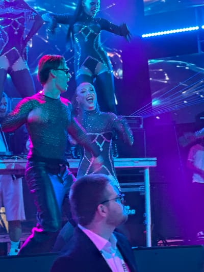 The image depicts a stage performance in the city of Tsilkani, Georgia. In the foreground, a man wearing glasses and a black shirt with a textured pattern and black leather pants is looking to the right with his mouth open. Behind him and to his right, a woman in a sparkling, dark outfit with a high neckline is laughing. Two other performers, one in a similar sparkling outfit and the other in a checkered-patterned outfit, are visible in the background. The stage is lit with vibrant blue and purple lights, and laser beams are projected across the scene. Stage equipment, including speakers and lighting fixtures, are visible. The overall atmosphere suggests a lively and energetic performance.