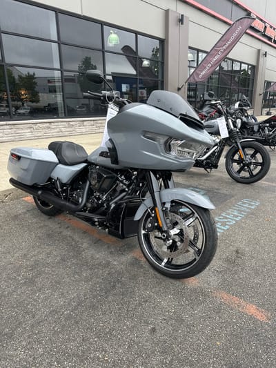 A grey Harley-Davidson motorcycle is parked in front of a building with large windows. The motorcycle is angled towards the viewer, with its front wheel turned slightly to the left. The motorcycle has a large front fairing, saddlebags, and a black exhaust system. In the background, other motorcycles are visible, as well as a banner that reads "BARNES HARLEY-DAVIDSON". The ground is paved with asphalt, and there are orange markings on the ground. The building appears to be a dealership or showroom. The scene is outdoors, and the lighting suggests it is daytime.