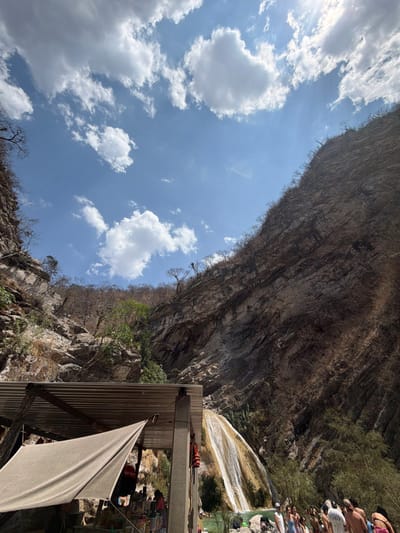 This image depicts a natural canyon environment near San Miguel Atlapulco, Mexico. The upper portion of the frame is dominated by a bright blue sky with scattered white cumulus clouds; the sun is visible in the upper right quadrant, partially obscured by a cloud. Steep, rugged rock faces define the canyon walls, extending from the mid-ground upwards. These cliffs display layered geological formations and are sparsely covered with dry, scrubby vegetation, including bare trees and bushes.
In the mid-ground on the right, a multi-stream waterfall cascades down an inclined, light-colored rock face into turquoise-green pools situated at the base of the canyon. A crowd of approximately 25-35 individuals is dispersed around and within these pools in the lower right of the frame, engaged in recreational water activities, dressed in swimwear or light attire.
In the lower left foreground, a partial man-made structure is visible. It features a corrugated metal roof supported by dark vertical pillars. Beneath this roof, a light-colored canvas tarp is stretched, partially obscuring the interior, where various unspecified items are present. A red and orange life vest hangs from one of the visible pillars.