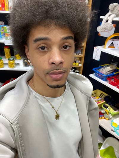 An adult male individual is depicted from the upper chest upward, facing the viewer directly in an indoor retail or storage setting in Plaisir, France. He has a dark afro hairstyle, a mustache, and a goatee, with medium skin tone. He wears a light grey hooded jacket over a matching t-shirt and a gold-toned chain necklace with an oval pendant depicting a tree of life design. A silver hoop earring is visible in his left ear. The background features dark walls with white shelving units. On the left, shelves hold multiple clear glass jars containing yellow items, alongside colorful packaged goods. On the right, shelves display a grey plush toy, a yellow plastic toy, blue and red plastic baskets filled with merchandise, a numerical chart affixed to the wall, a beige basket containing various colorful children's toys and books, and a white and green baby feeding chair.