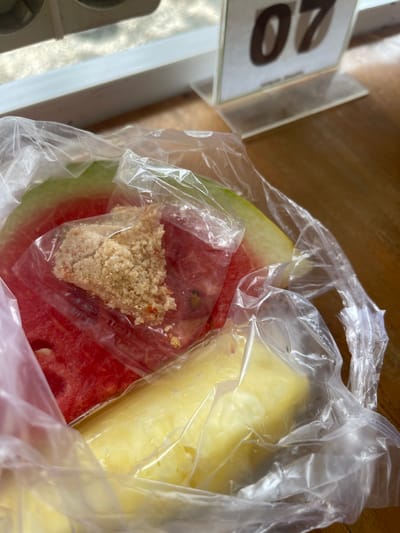 A close-up shot captures a plastic bag containing pieces of fruit, likely watermelon and pineapple, placed on a wooden table. A small portion of a numbered table marker, displaying "07", is visible in the background, suggesting a restaurant or dining establishment setting. The lighting indicates an indoor environment, possibly with natural light filtering in. The scene appears to be within the city of Pulokiton, Indonesia.