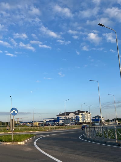 This media file captures an outdoor scene in a modern setting, likely during the daytime under a clear blue sky with scattered white clouds. The perspective is from a roadside, looking towards a row of contemporary buildings with blue and white facades. These structures appear to be multi-story and arranged along a street or thoroughfare.
Several streetlights with a distinctive curved design are visible, spaced along the roadside and stretching up into the sky. A road with lane markings, including a prominent white curved line, occupies the right side of the frame. Adjacent to the road is a grassy area bordered by a metal fence or railing. Parked cars are visible in front of the buildings, suggesting a commercial or residential area.
In the distance, a construction crane can be seen, hinting at ongoing development in the vicinity. A round blue and white traffic sign with two opposing arrows, indicating a potential change in road direction or lane usage, is positioned on a pole. The overall atmosphere is bright and open, indicative of fair weather. The presence of the moon in the sky, though small and faintly visible, suggests it might be late afternoon or early morning. No individuals are clearly discernible in the image. The location context provided is Холмогоровка, Russia.