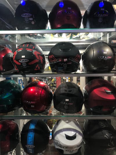 The image shows a shelf display of various motorcycle helmets in a retail setting. The helmets are arranged in rows on glass shelves. The helmets vary in color and design, with some being plain black, red, or green, while others feature more intricate graphics and patterns. Some helmets have logos and labels visible, such as "u.p.Co.", "SIS", and "A.B.S. REINFORCED". There are also blue LED lights shining on some of the helmets at the top of the display. The background is blurred but suggests a shop interior with more merchandise or shelving. The overall impression is that of a helmet shop or a section of a larger store selling motorcycle accessories. This scene is located in Karandugoda, Sri Lanka.