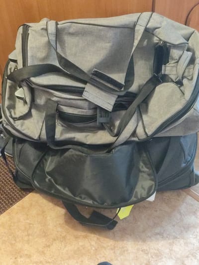Two duffel bags are stacked on a floor. The top bag is grey, and the bottom bag is black. The grey bag has a black strap that runs diagonally across it. The black bag has a large pocket on the front. The floor is tiled with a light brown and tan pattern. The background is a wooden wall. The image is taken from a high angle. There is no indication of the location in the city of Askiz, Russia.