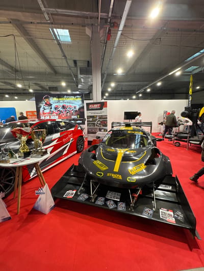 The scene depicts an indoor exhibition within a large hall, located in Wolica, Poland. The floor is covered entirely with a red carpet. The ceiling is high, featuring exposed metal trusses, industrial lighting fixtures, and ventilation ducts.

In the mid-ground, two distinct race cars are on display. To the left, a red and black hatchback-style race car is presented, featuring abstract white and grey graphics and "KUBIT MOTORSPORT" branding. Adjacent to this car, a small, round white table with light wooden legs holds several golden trophies and various smaller awards. To the right, a dark grey/black open-wheel or highly modified sports car, identified with "KUBIT MOTORSPORT" and "LOTUS EXIGE" branding in yellow lettering, is positioned with a prominent front aerodynamic package, including a large diffuser-wing assembly. Several small informational cards are placed on this front diffuser.

In the background, a large banner features an image of a motorcyclist and text including "DRUŻYNOWY PUCHAR ŚWIATA" (Team World Cup) and the date "29.08.2026." Next to it, a "KUBIT MOTORSPORT" stand displays "PROFESJONALNY SPRZĘT WYŚCIGOWY" (Professional Racing Equipment) and images of automotive parts. Several individuals are present, observing the exhibits. One person in a white shirt stands behind the dark grey race car, near a black display screen. Other people are visible moving or standing in the background. The overall environment suggests a motorsport or automotive trade show.