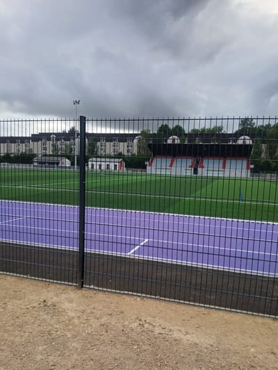 The image shows an outdoor sports facility with a fence in the foreground. The facility has a green artificial turf field next to a purple track with white lines. In the background, there are buildings with multiple stories, some trees, and a small grandstand with red seats. A lamppost with floodlights is visible in the distance, suggesting it could be used for evening events. The sky is overcast with grey clouds, indicating a cloudy day. The ground next to the track appears to be dirt or gravel. There is no visible text in the image. The scene depicts a sports complex, possibly a stadium or training ground, in Charleville-Mézières, France. The overall mood is neutral, focusing on the architectural and sporting elements of the location.